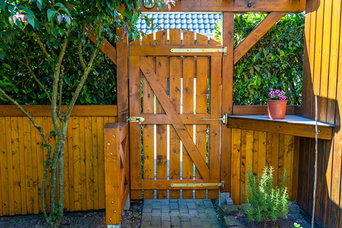 A wooden garden gate with a flower pot on a ledge, surrounded by lush greenery and wooden fencing.