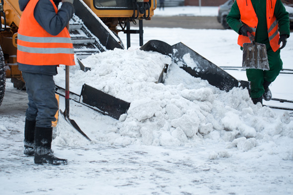 Workers removing snow with shovels and a snowplow machine in a winter setting.