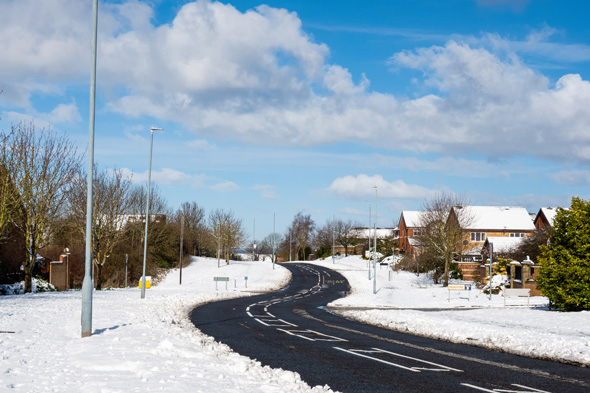 A snowy residential street under a blue sky, featuring trees and houses alongside a winding road.