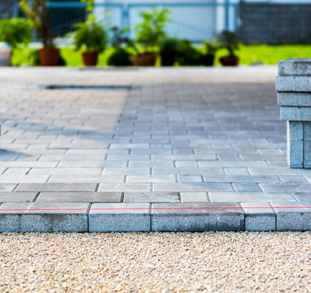 Paved walkway with gray blocks and a red outline, surrounded by green plants and gravel. Ideal for outdoor landscaping.