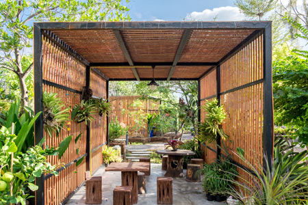 A rustic outdoor seating area featuring wooden benches and tables surrounded by lush greenery and decorative plants. The structure has a bamboo design with open sides, creating a serene and inviting atmosphere. Ideal for relaxation or gatherings in nature.